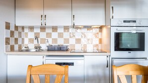 Una cucina con mobili bianchi, zoccolo a scacchi, lavello, lavastoviglie e forno. Sono presenti due sedie di legno attorno a un tavolo.