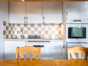 Cocina con armarios blancos, salpicadero de cuadros, un lavaplatos, lavamanos y horno. Hay dos sillas de madera junto a una mesa.