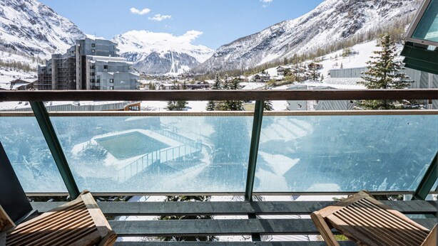 Un balcone con ringhiera in vetro, sedie di legno e vista sulle montagne innevate e sugli edifici.