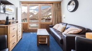Un soggiorno moderno con una scrivania in legno, un divano in pelle, una televisione e un'ampia finestra vista montagna.