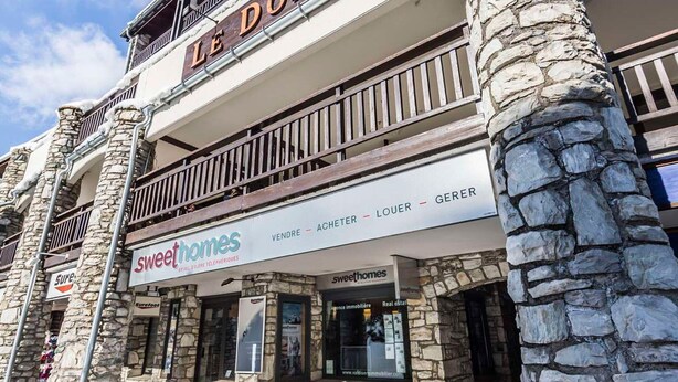 A two-story building with a stone facade and a balcony. The ground floor has a sign for 'Sweethomes' with a 'VENDRE - ACHETER - LOUER - GERER' sign above it. - Comfortable studio in center, balcony, 2-3 pers. (Val-d'Isère)