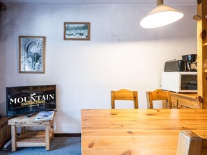 A room with a wooden dining table, chairs, a television, and a kitchen area with a stove and fan.