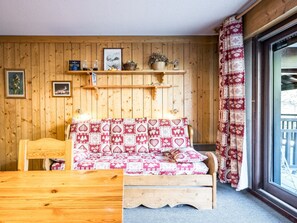 A wooden cabin interior with a bed, a wooden table, a shelf with various items, and a window with curtains.