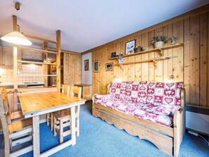 A wooden interior with a dining table, chairs, a sofa with patterned cushions, and a wall-mounted shelf with decorative items.