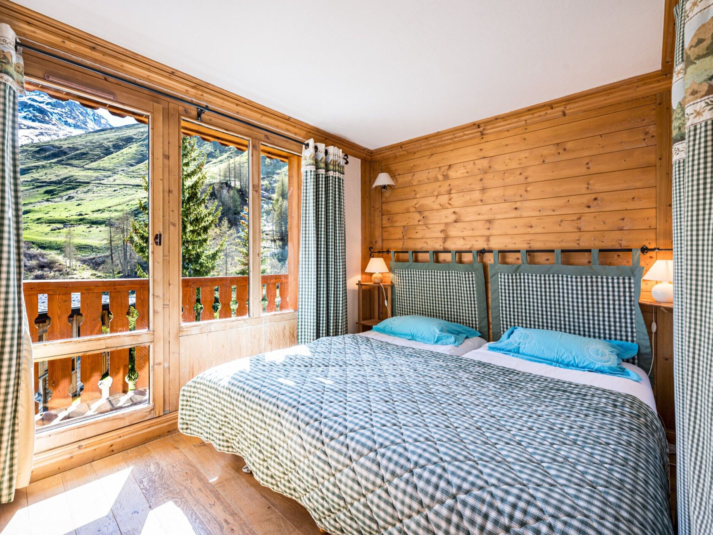A bedroom with a large window, wooden walls, a bed with checkered bedding, and two bedside lamps.