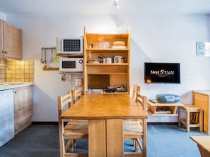 Uma cozinha compacta com mesa de madeira, cadeiras, micro-ondas, máquina de café e televisão na parede.