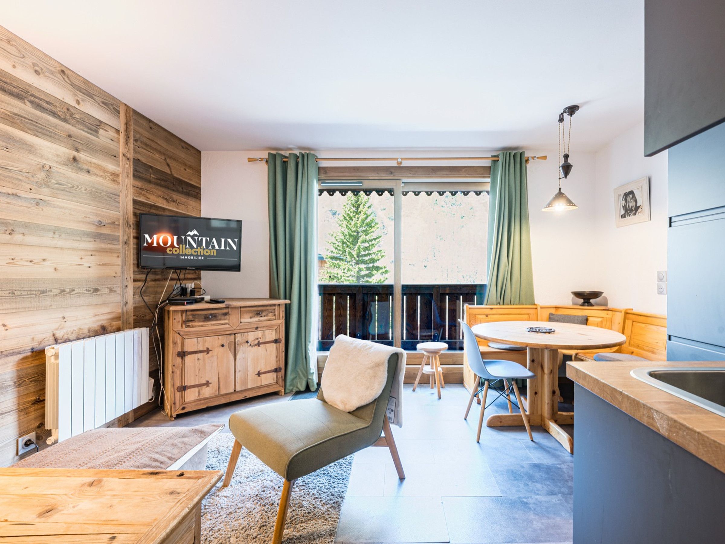 Eine moderne Küche mit Holzschränken, ein Essbereich mit Tisch und Stühlen, ein an der Wand befestigtes Fernsehgerät und Blick ins Freie.