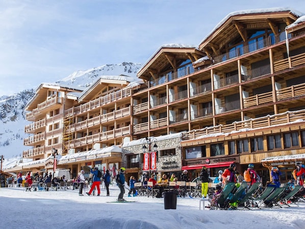 A multi-story wooden building with balconies, ski equipment, and a snow-covered mountain in the background.