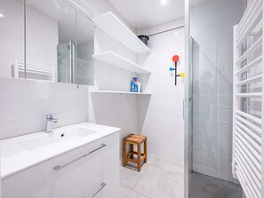 Un bagno moderno con lavandino, specchio e doccia bianchi. Sopra lo specchio ci sono degli scaffali e un piccolo sgabello di legno.
