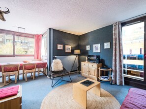A room with a dining table, chairs, a sofa, a chair with a fluffy cushion, a small wooden cabinet, a lamp, and a radiator.