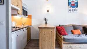 A compact kitchen with wooden cabinets, a microwave, and a sink. There's a wooden table and a sofa with cushions. A poster is on the wall.