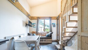 A room with a wooden dining table, white chairs, a sofa with cushions, a window with a view of mountains, and a wooden staircase. - Renovated Studio with Mezzanine – Val-d'Isère Center (Val-d'Isère)