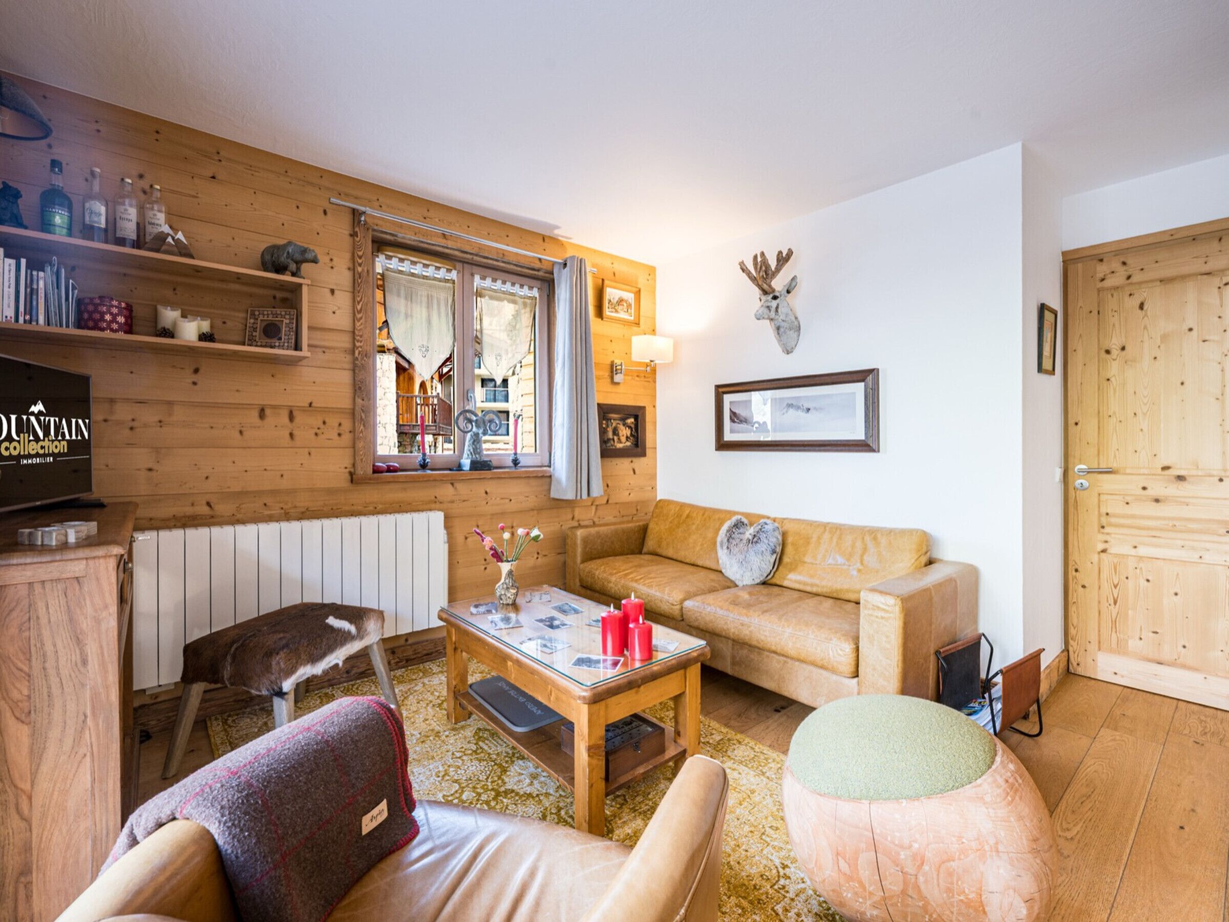 A cozy living room with a sofa, armchair, and coffee table, wooden walls, a mounted deer head, and a window with curtains.