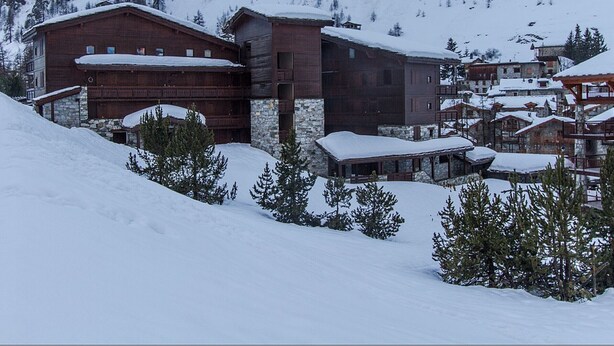 A snow-covered mountain lodge with multiple wooden buildings, including a large garage and several smaller structures.
