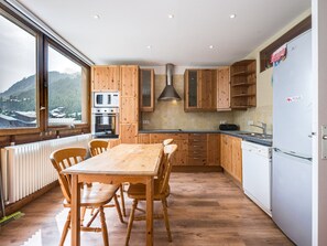 Uma cozinha com armários de madeira, uma mesa de jantar com cadeiras, forno embutido e vista para uma paisagem montanhosa por uma grande janela.