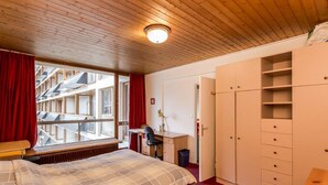A bedroom with a wooden ceiling, a bed with a plaid duvet, a desk with a chair, a wardrobe, and a window with red curtains. - Comfortable 3-room apt · Near center · 5 min from slopes · Parking · Wifi (Val-d'Isère)