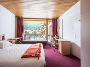 A hotel room with a bed, a desk, a chair, and a view of a village through large windows.