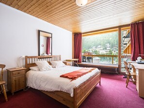 A bedroom with a wooden bed, a nightstand, a desk, a chair, a window with a view of a village, and a red carpet.