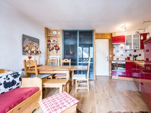 A cozy room with a wooden bed, a dining table with chairs, a kitchen area with red cabinets, and a large window. - Cozy 2-room apartment in La Daille, 4 people, ski-in/ski-out (Val-d'Isère)