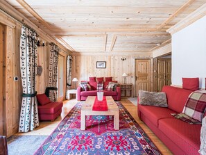 A cozy room with wooden walls and ceiling, a red sofa, a matching armchair, a coffee table, and a patterned rug.