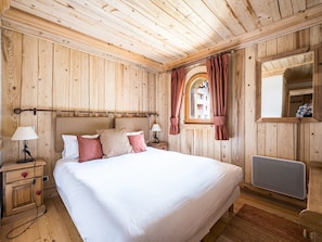 Una habitación de madera con una cama, dos mesitas de noche, una ventana con cortinas, un radiador y un espejo.
