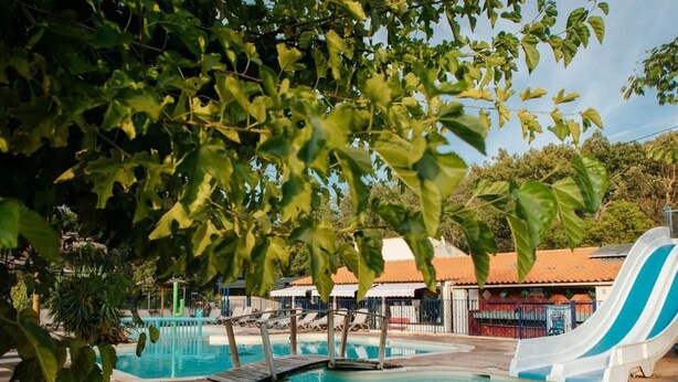 Une piscine avec un toboggan, entourée d’arbres et bordée d’un bâtiment au toit rouge.