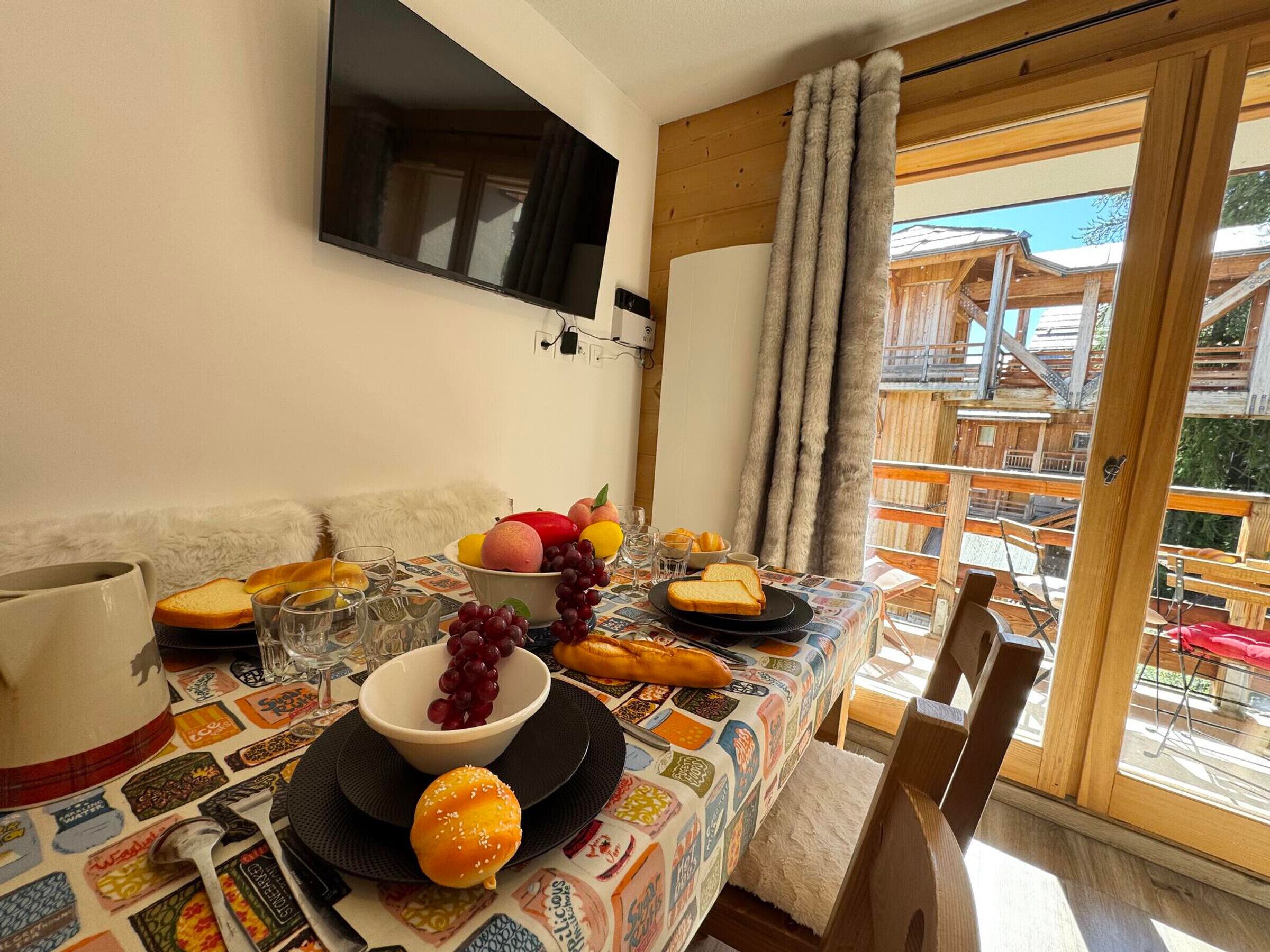 Una mesa de comedor dispuesta con comida y bebidas, un televisor montado en la pared y vistas al exterior.