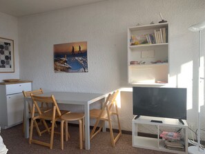 Quarto com escrivaninha branca, duas cadeiras de madeira, televisão sobre um suporte, estante e quadro na parede.