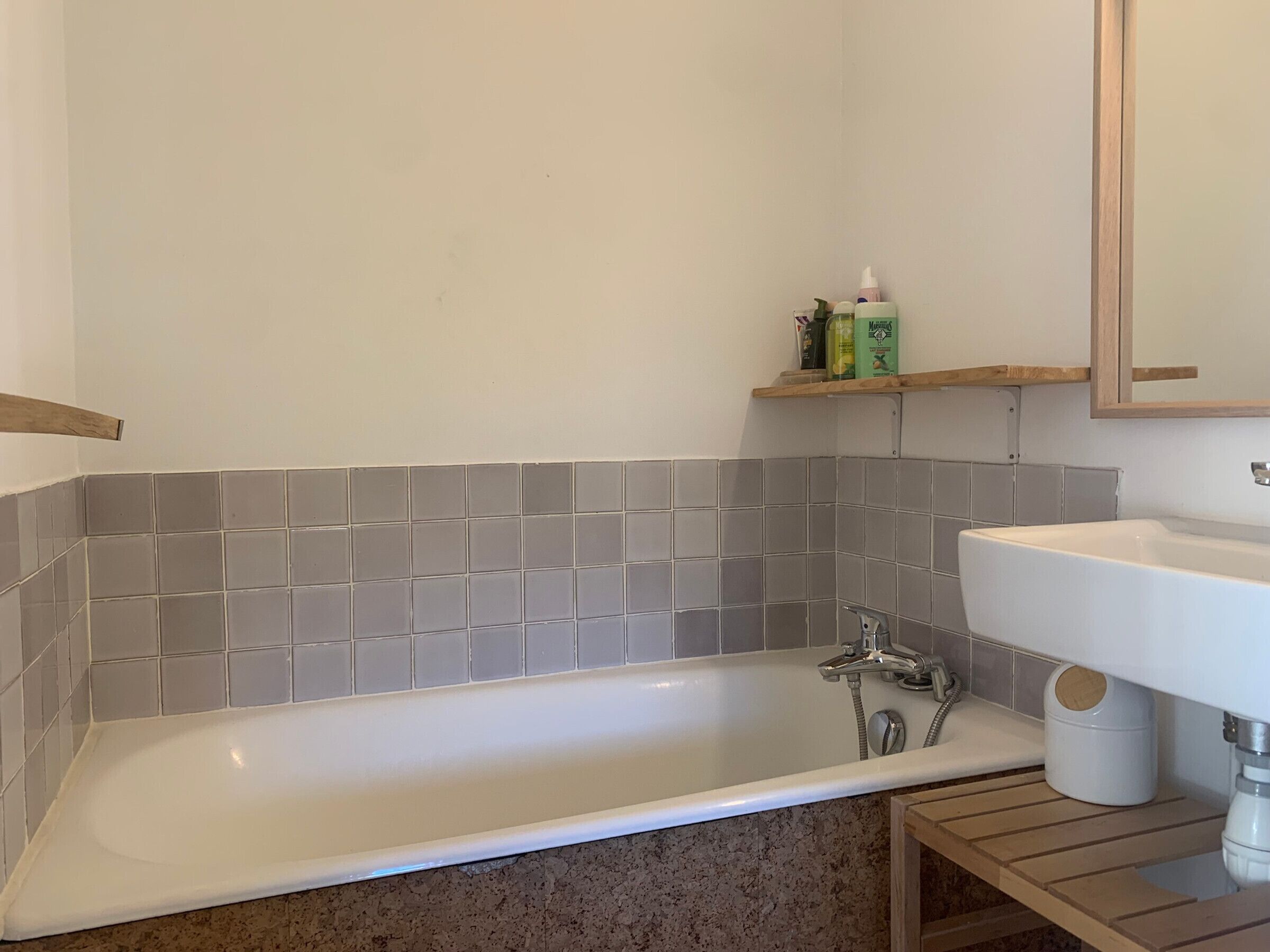 Een badkamer met een witte badkuip, grijze betegelde muren, een houten plank met toiletartikelen en een wit wastafel.