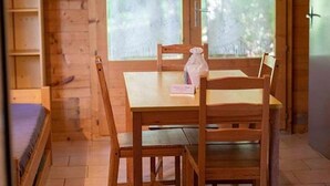 A wooden interior with a table and chairs, a window with a view of greenery, and a shelf with items.