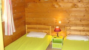 A wooden cabin room with two beds, each with a green bedspread and white pillows. There are two wall-mounted lamps with green shades.