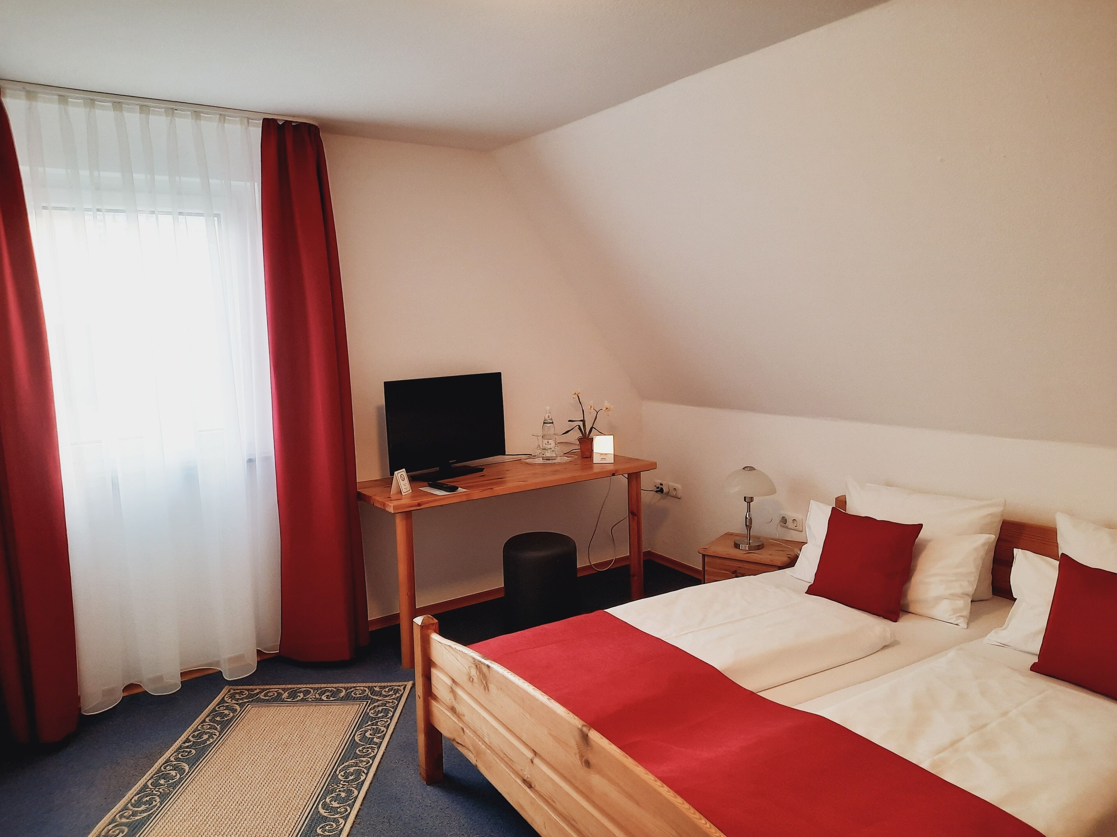 Habitación de hotel con una cama, un escritorio con televisor, una mesita con lámpara y una ventana con cortinas.