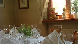 Une table à manger dressée avec des verres à vin et une fourchette, un ours en peluche sur une étagère, et une fenêtre avec des rideaux.