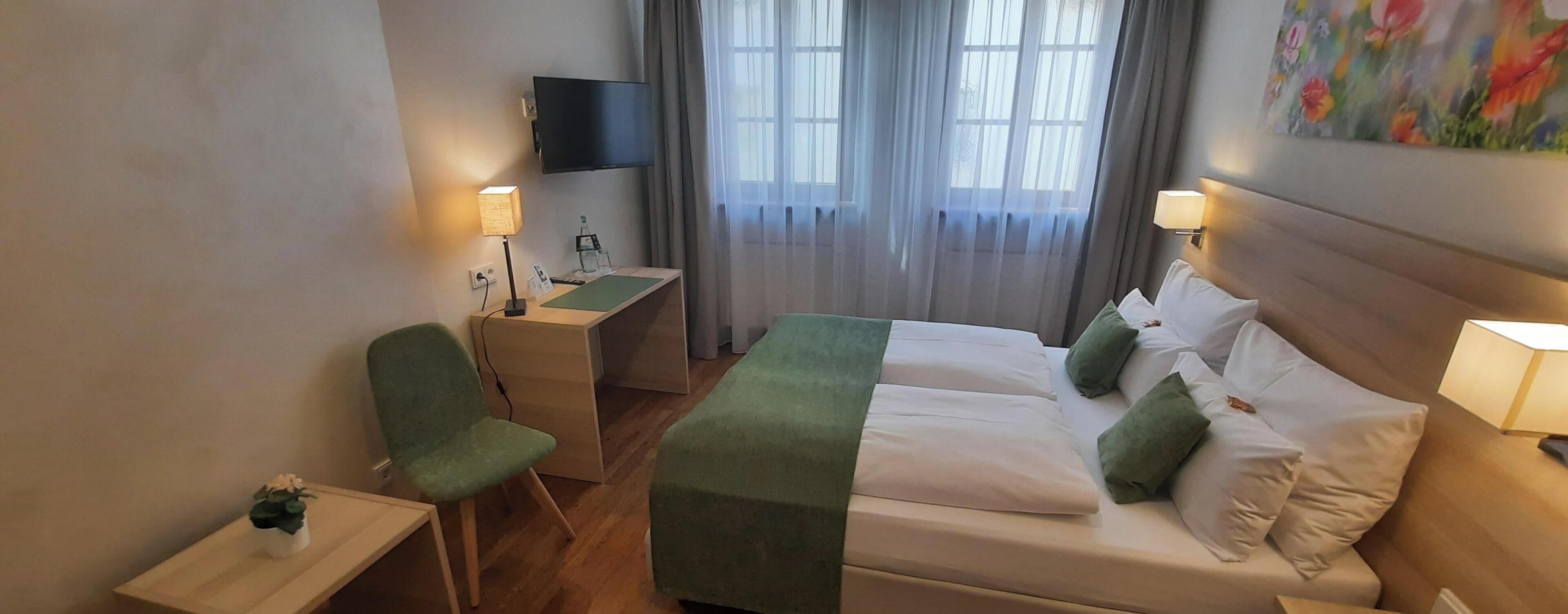 A hotel room with a bed, a desk, a chair, a TV, and a painting on the wall.