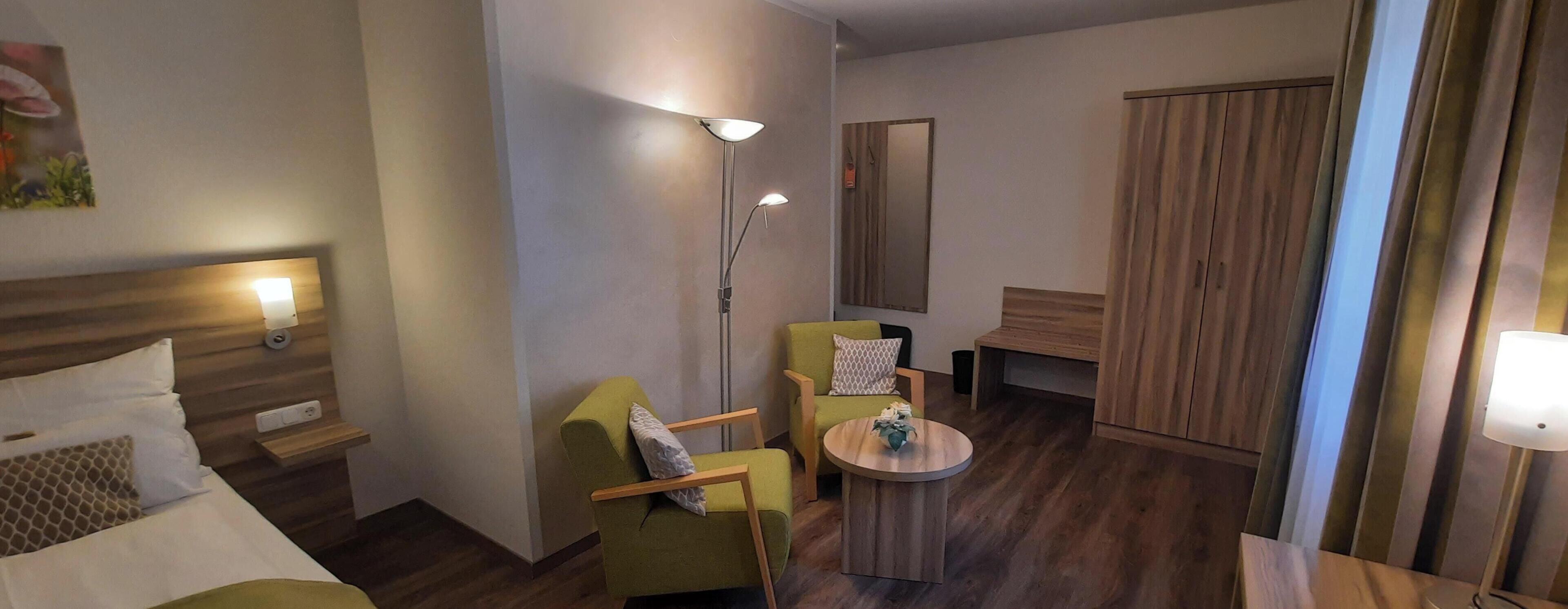 Een hotelkamer met een bed, twee groene fauteuils, een kleine ronde tafel, een vloerlamp en een kledingkast.