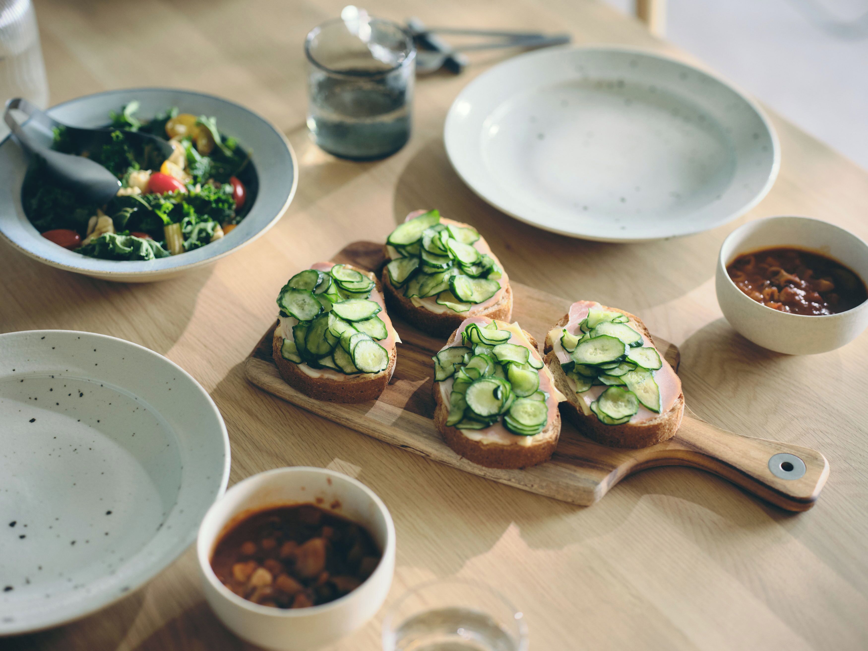 Una mesa de madera con tres sándwiches abiertos cubiertos de rodajas de pepino, un bol de ensalada, un bol de sopa y un vaso de agua.