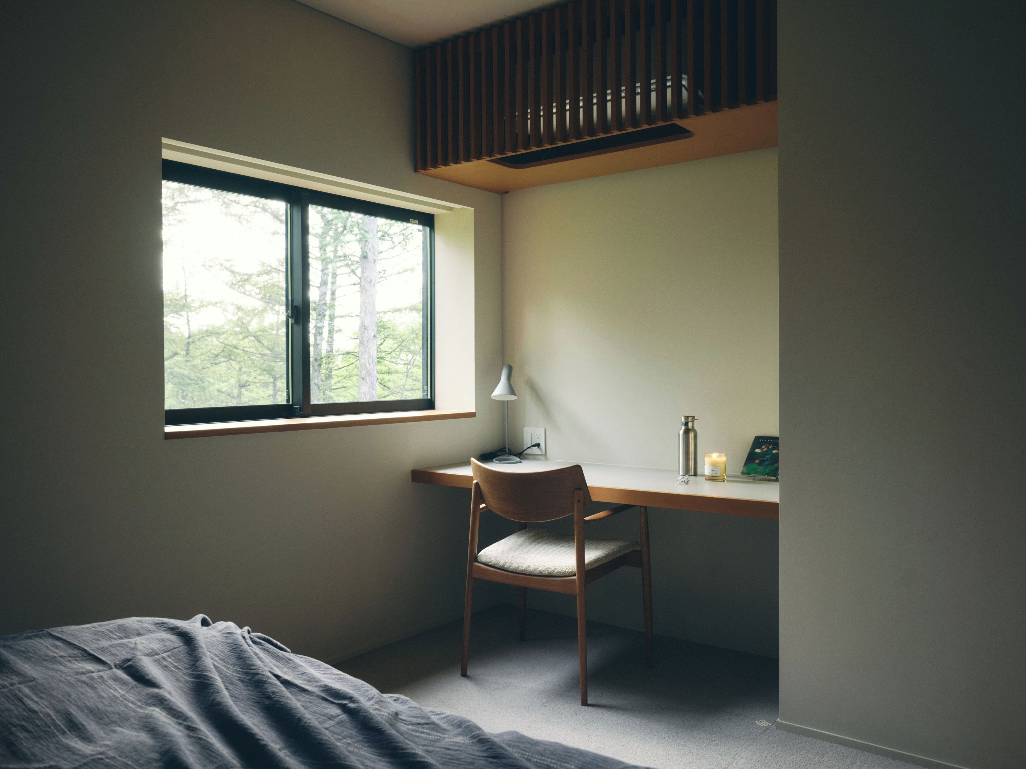 A bedroom with a bed, a desk with a chair, a window with a view of trees, and a bedside table with a lamp and a candle.