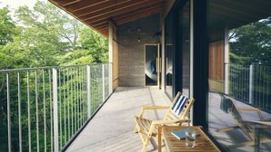 Un balcon doté d’un plancher en bois, de deux chaises en bois, d’une table avec un verre et un livre, et offrant une vue sur les arbres à travers des barreaux en métal.