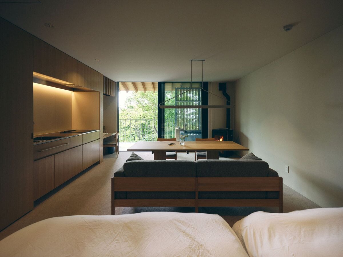 A modern, minimalist bedroom with a large bed, a wooden bench, and a dining area with a table and chairs.