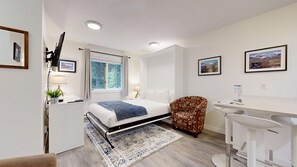 A hotel room with a bed, a desk, a chair, a window with a view of trees, and a picture on the wall. - Snow Creek #39 Suite | 3 Peaks Rentals (Sun Peaks)