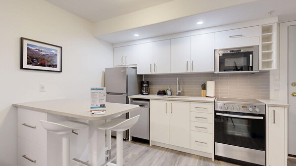 A compact kitchen with white cabinetry, a built-in microwave, a stove, and a coffee maker. - Snow Creek #39 Suite | 3 Peaks Rentals (Sun Peaks)
