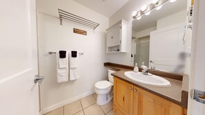Ein Badezimmer mit einem weißen Waschbecken, Holzschränken, einer Toilette, einem Handtuchhalter mit zwei Handtüchern, einem beleuchteten Spiegel und einem Schild über dem Handtuchhalter.