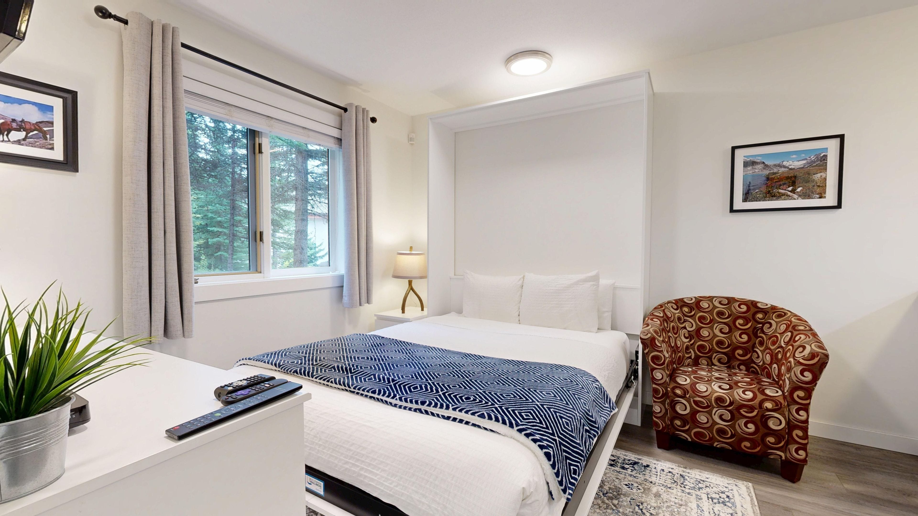 A hotel room with a bed, a desk with a plant, a chair, a lamp, and a window with curtains.
