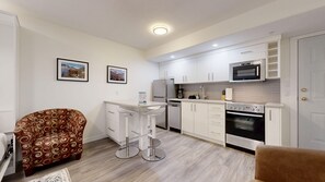 Cocina compacta con armarios blancos, microondas, refrigerador y estufa. Hay un taburete de bar y un sillón estampado.