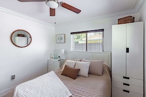 A bedroom with a bed, a bedside table, a mirror, a window with blinds, a ceiling fan, and a wardrobe.