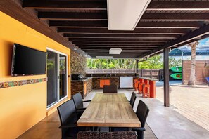 An outdoor patio with a wooden table and chairs, a TV mounted on the wall, and a stone backsplash. - Waterfront 3BR with Pool, Tiki Hut & Mini Golf (Miami Gardens)