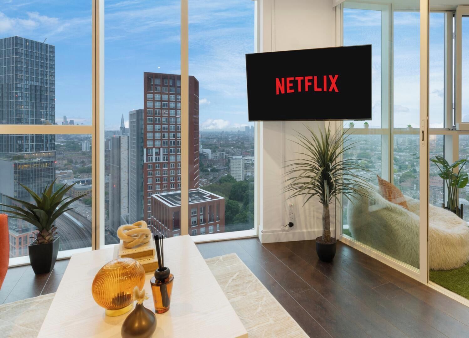 Ein modernes Wohnzimmer mit großem Fenster, einem Flachbildfernseher, der 'NETFLIX' zeigt, und Blick auf die Stadtlandschaft.