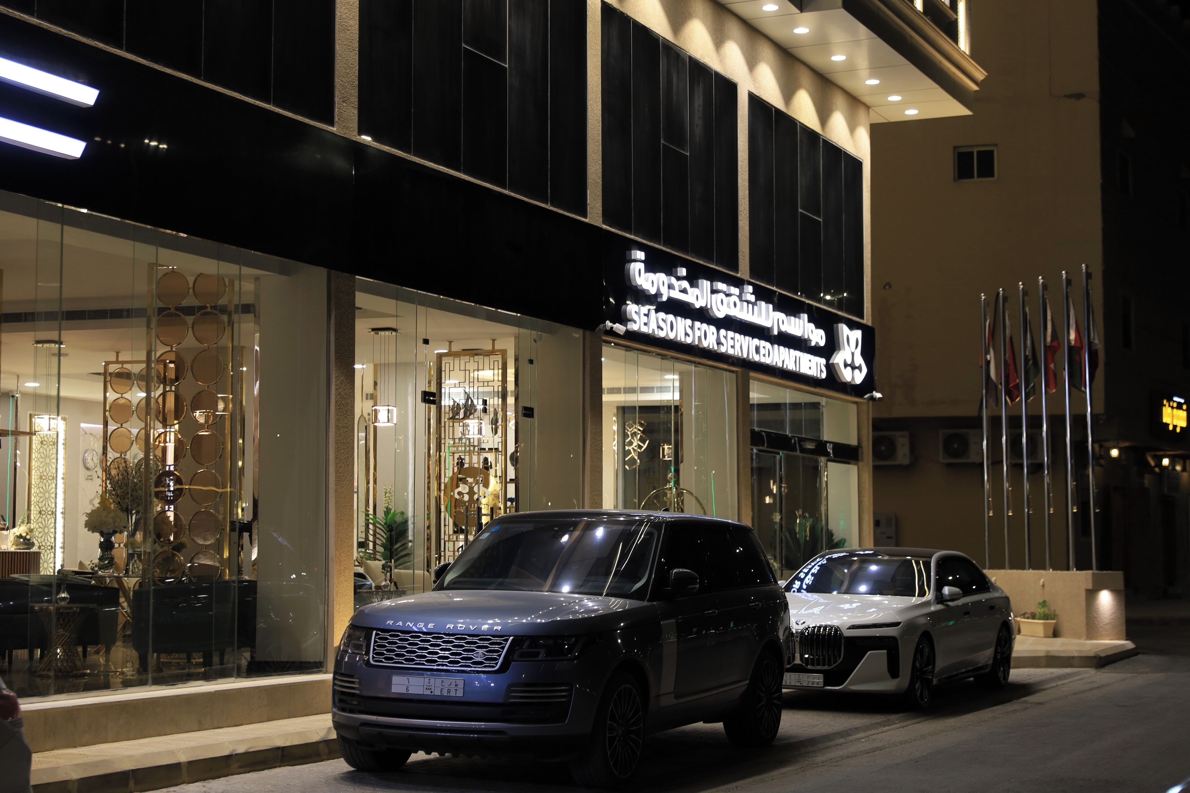 A modern hotel with a glass facade, a sign indicating it offers seasonal services, and two luxury cars parked in front.