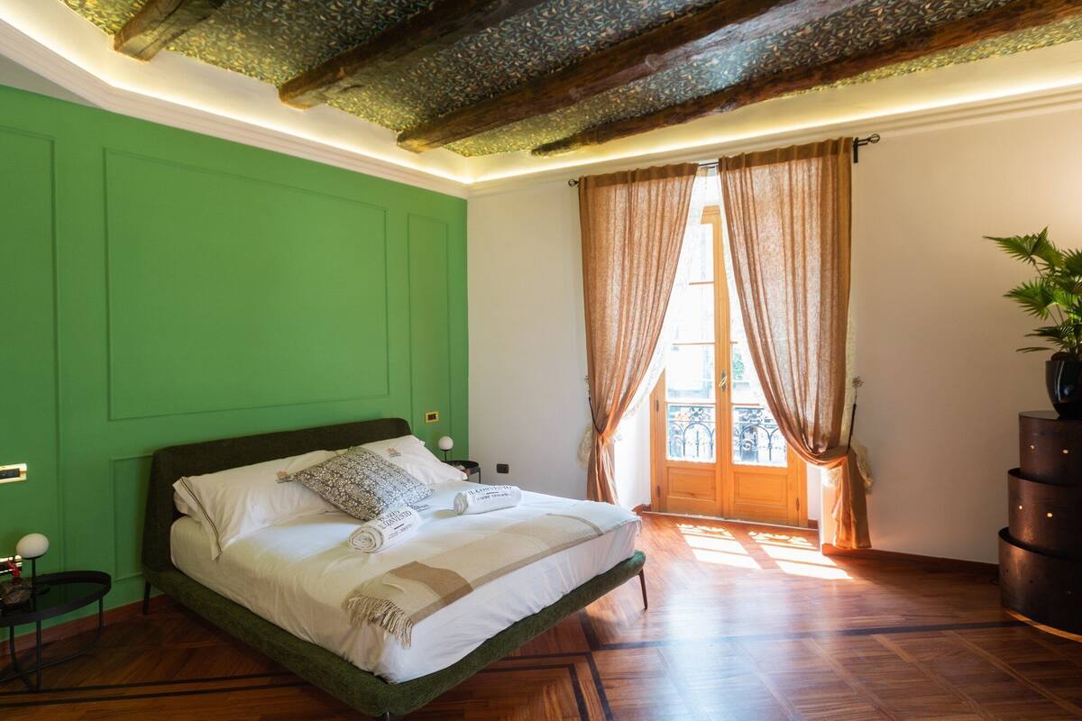 Un dormitorio con pared verde, suelo de madera, cama con ropa de cama blanca, mesita de noche, planta en maceta y ventana con cortinas.