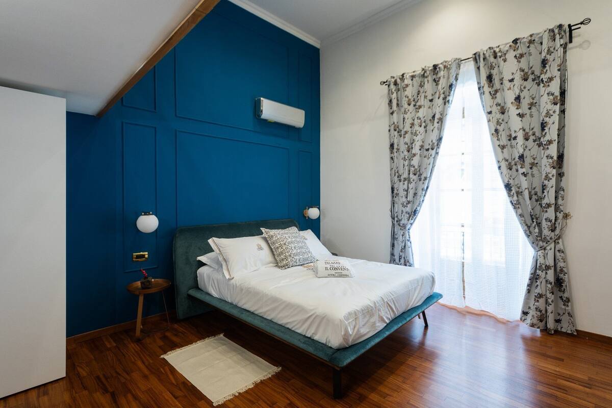 Un dormitorio con pared azul, una cama con ropa de cama blanca, suelo de madera y una ventana con cortinas estampadas.